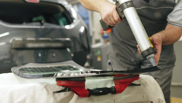 Process of replacing automobile rear window. Medium closeup shot of caucasian mechanic in grey uniform holding specialistic glue gun and applying glue to separated glass. High quality 4k footage
