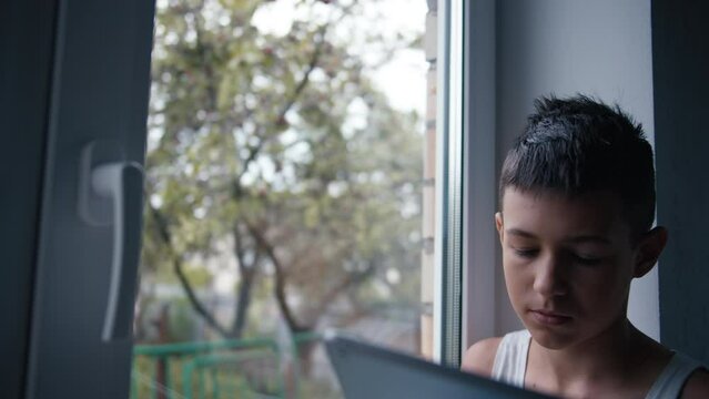 serious teen boy sits near the window and uses the tablet in autumn weather, cinematic steadicam shot