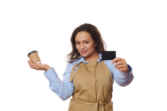 Attractive waitress bartender, barkeeper in beige chef apron, smiles looking at camera, holds a cardboard cup of hot drink to go and plastic bank card, isolated over white background. Food and drink