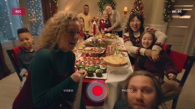 Caucasian woman with selfie stick takes family group photo. Multi cultural family celebrating Christmas or New Year 2023. Table with dishes. Atmosphere of family Christmas dinner at home. Camera view.