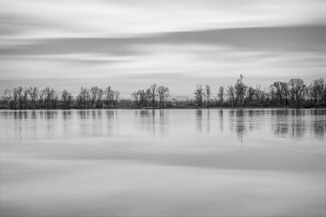Fototapeta premium View across a calm river at a treed island, sky and trees reflected in water, nobody