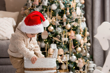 A beautiful little boy in a Santa Claus hat is sitting in a basket and laughing merrily against the background of a Christmas tree. A New Year's gift. The concept of celebrating Christmas.