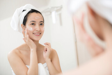 Happy young Asian woman applying face lotions while wearing a towel and touching her face in bathroom