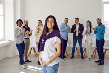 Fototapeta premium Portrait of confident happy young woman in casual clothes against background of other people. Office worker or company manager working in business center posing against backdrop of applauding people.