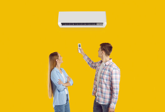 Happy Married Couple Turn On Air Conditioner Isolated On Solid Yellow Color Background. Young Husband And Wife Using Remote Control To Adjust Temperature On AC Unit On Wall. Air Conditioning Concept