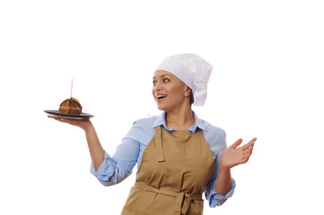 Isolated on white background, female chef confectioner in beige apron, smiling while looking at a piece of homemade chocolate cake with candle on a plate in her hands. Baking for anniversary, birthday