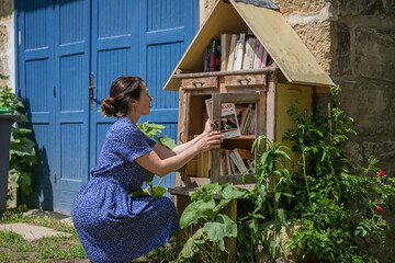 Woman takes the free books on the street. Pretty girl choosing a book to borrow in street library. Sidewalk Library box for public in Residential Neighborhood - obrazy, fototapety, plakaty