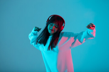 Young Asian woman wearing wireless headphones listening music and dancing in neon light background. Emotions and ad concept..