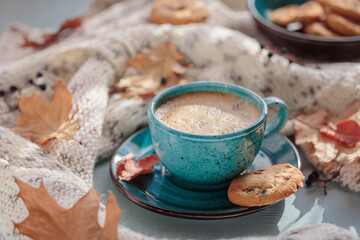 Autumnal composition with white knitted scarf, blue cup of coffee, cookies and dry yellow leaves on a table. Autumn mood.