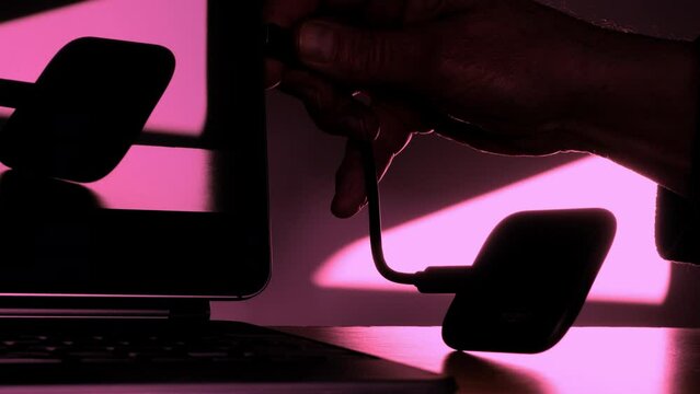 Closeup in near silhouette of a man&rsquo;s hand connecting the cord / cable of a small, lightweight, portable external SSD hard drive, to a laptop computer.