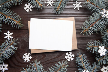 Christmas card mockup with envelope and green fir tree branches on brown wooden background, top view, flat lay