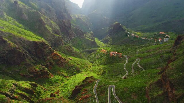 Winding road in mountains green landscape and glowing horizon during sunrise with sun rays. Masca Gorge and village. New asphalt paved smooth highway freeway, countryside with cars. Springtime nature.