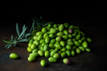 Pile of fresh green olives and branch with leaves on black background. Dark low key photo. Copy space. Blurred background. Selective focus.