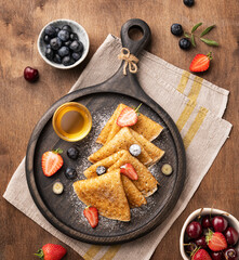 Homemade crepes with fresh berries and honey on a dark wooden plate on a brown background.