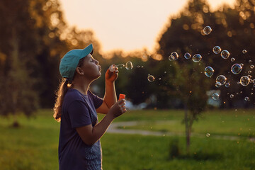 A girl of 10-12 years old blows soap bubbles in the park at sunset. School-age child enthusiastic...