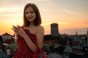 Young beautiful female model in the red dress enjoy the glass of wine on the roof of Old city of...