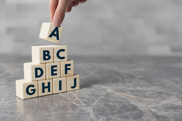 The hand arranges wooden blocks with the alphabet from A to J by putting a block with the letter A...