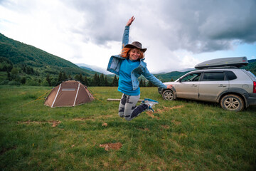 Woman jumping for happiness, having fun and dancing on the top of the mountain. Concept of travel and wandering, tent and camping. Off road vehicle