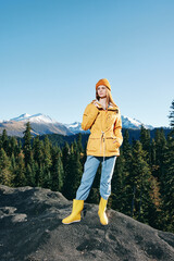 Naklejka premium Young woman with red hair full-length hiker in yellow raincoat and cap runs and walks on a mountain trip in the fall and hiking in the mountains at sunset freedom