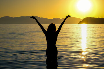 View of a woman standing in the sea  with her hands up in the sky enjoying the amazing sunset at...