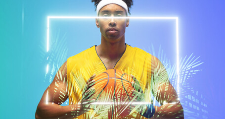Naklejka premium Portrait of biracial basketball player holding ball in front of illuminated rectangle and plants