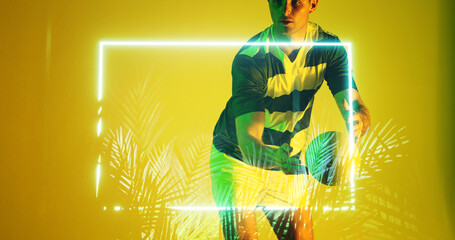 Caucasian rugby player catching ball by illuminated rectangle and plants over yellow background