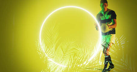 Caucasian male player catching rugby ball by illuminated circle and plants on yellow background