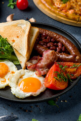 English Breakfast with two eggs, sausage, bacon, toasts and vegetables, red beans on a black plate vertical