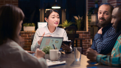 Asian woman speaking at table in business meeting, corporate presentation discussion. Coworkers brainstorming, marketing strategy planning, research analytics, medium shot. Tripod shot.