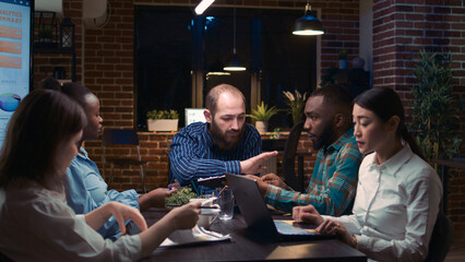 Company team group brainstorming in business meeting, discussing presentation, diverse coworkers communication. Employees planning sales strategy in coworking space at night time