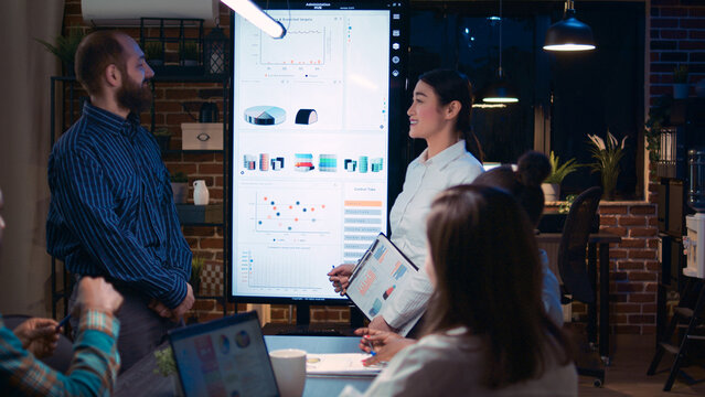 Diverse coworkers presenting company annual investment profit report, employee planning sales strategy in coworking space. Office workers in business meeting, data graphs on digital board