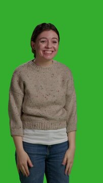 Vertical video: Front view of close up young adult greeting person with wave, smiling in studio. Relaxed casual woman waving hi or hello on camera, standing over full body green screen background and