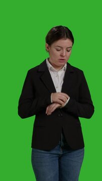 Vertical video: Front view close up of impatient office worker checking time on wristwatch, standing over isolated greenscreen backdrop. Young woman running late and looking at clock, waiting for