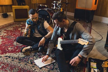 Male guitarists of a music band learning a new song, sitting on the floor with notes. High quality...