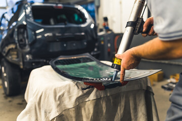Installing process of car window. Indoor shot of professional unrecognizable mechanic using polyurethane glue gun. Black blurred car in the background. High quality photo - obrazy, fototapety, plakaty