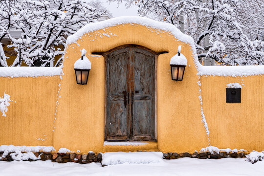 Snowy scene in Santa Fe of adobe wall, glowing lights and rustic door