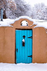 Winter scene of snow-covered adobe wall with a turquoise colored door and red chile ristra in Santa...