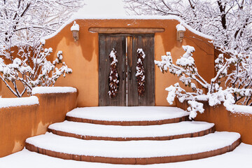 Winter scene of snow-covered adobe wall with rustic wood doors and chile ristras in Santa Fe, New...