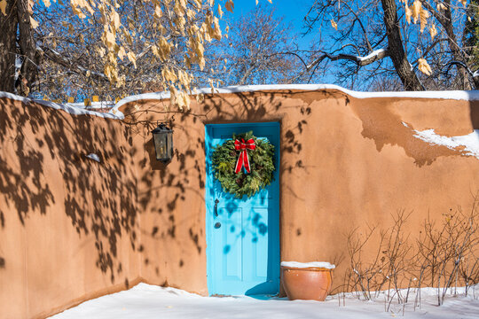 Snowy winter scene of Christmas wreath on turquoise colored door in adobe wall in Santa Fe, New Mexico