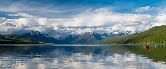 Panorama of snow-capped mountain peaks and red boat under dramatic sky reflected in the calm waters of Lake McDonald, Glacier National Park, Montana