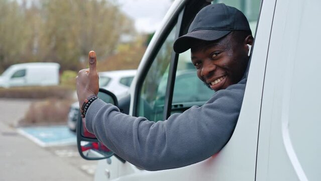 Black young adult delivery man sitting inside white van leaning out of the windo to look into camera smiling with thumbs up. High quality 4k footage