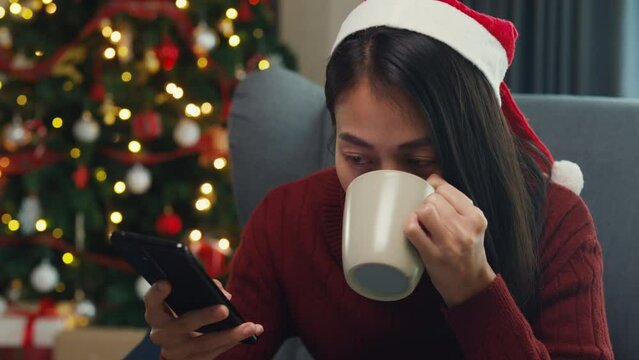 Positive Asia female wear red sweater new normal celebrate christmas smell aromatic coffee cup sit on wing chair check online greeting message from friend and family on phone in living room at house.