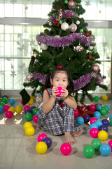 Fototapeta na wymiar little Asian girl sitting in front of a Christmas tree with colorful balls