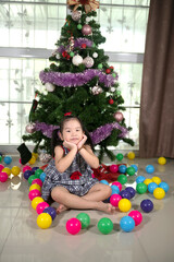 Fototapeta na wymiar little Asian girl sitting in front of a Christmas tree with colorful balls