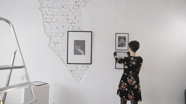 A woman gallery owner hangs new paintings and collages on the wall of the exhibition hall. Gallery with an exhibition of contemporary art.