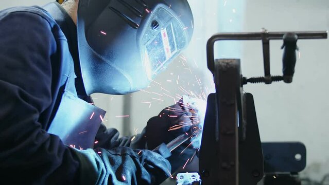 Welder in a mask welds metal parts fixed in a welding yews close up. Worker at an industrial site is engaged in welding work using personal protective equipment