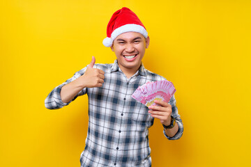 Fototapeta na wymiar Excited young Asian man 20s wearing a Christmas hat showing cash money rupiah and thumb up sign isolated on yellow background