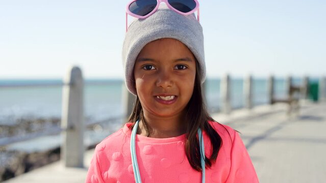 Child, face and fashion at beach promenade with a smile for travel, happiness and winter holiday at sea with a hat and pink clothes. Portrait of girl from Philippines happy about vacation and freedom