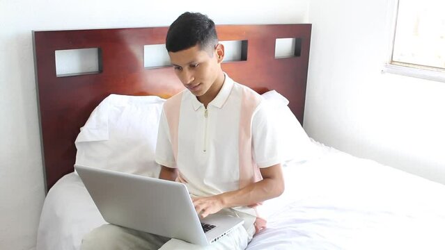 mexican young male student doing homework on laptop sitting on bed. hispanic man at home office