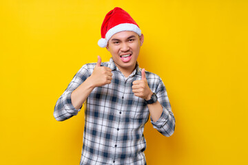 Smiling young Asian man wearing a plaid shirt in a Christmas hat posing showing thumb up like gesture on yellow background. celebration Christmas holiday and New Year concept